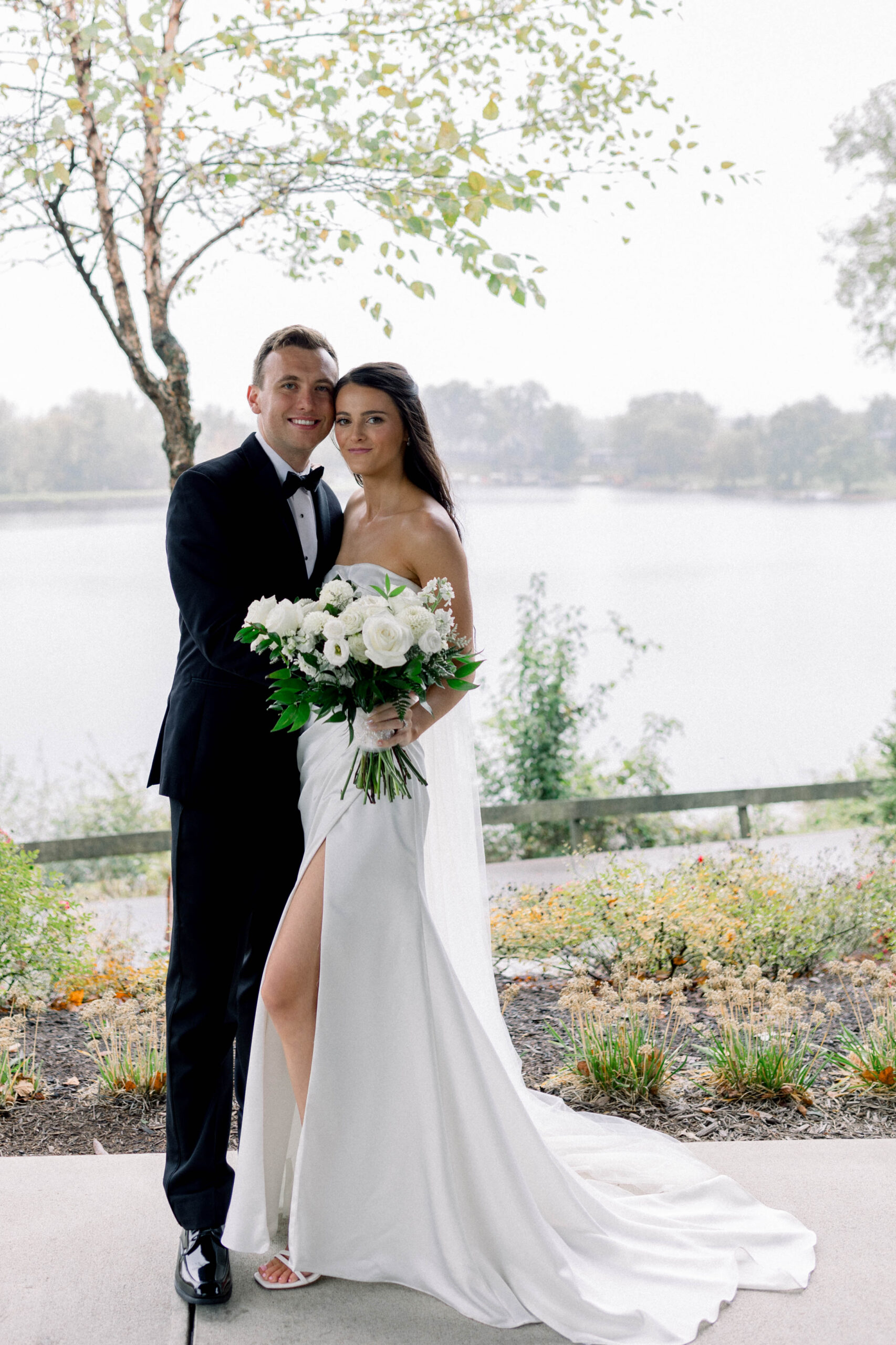 wedding couple in in a black tux and white dress with a rainy day lake behind them